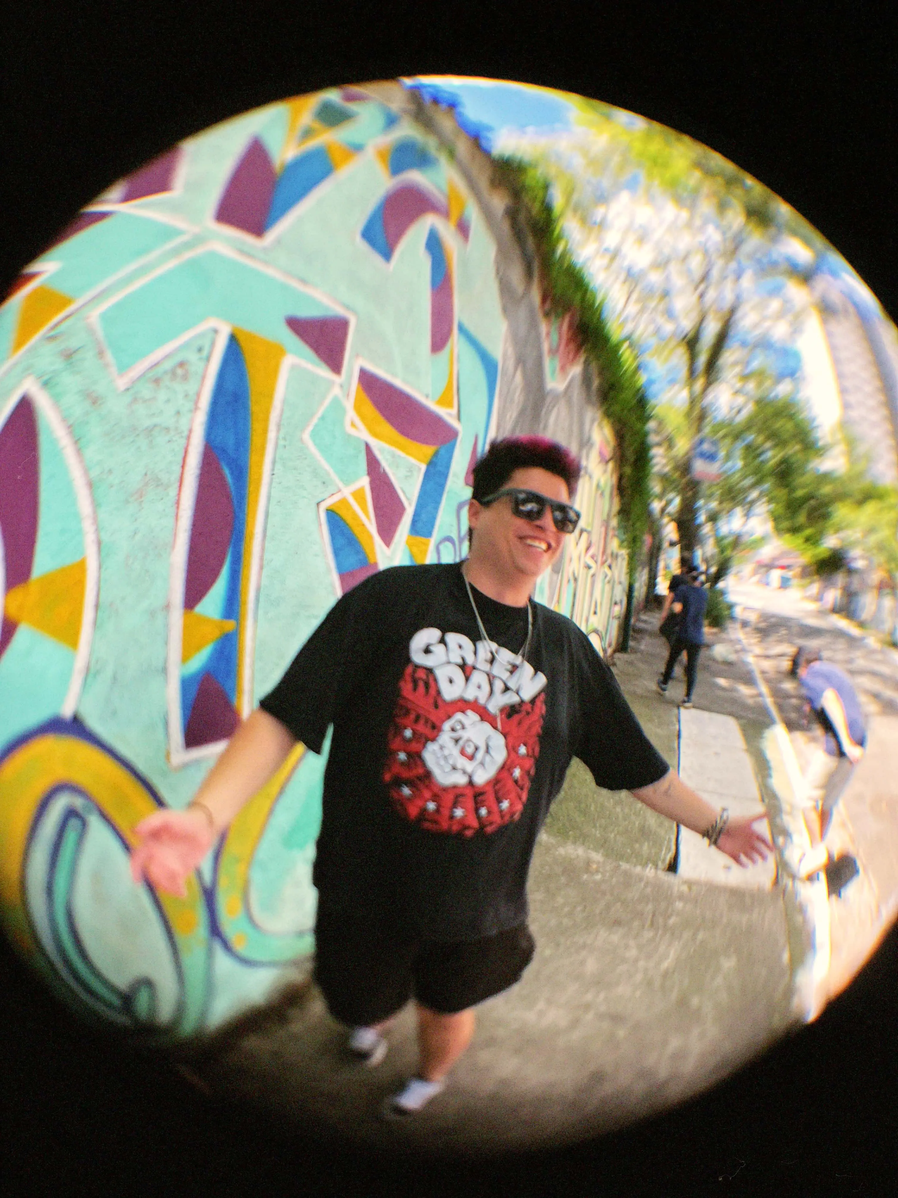 Guitarrista Tavinho está sorridente com óculos escuros e camiseta preta do Green Day. Ele está com os braços abertos em frente ao muro de grafite.